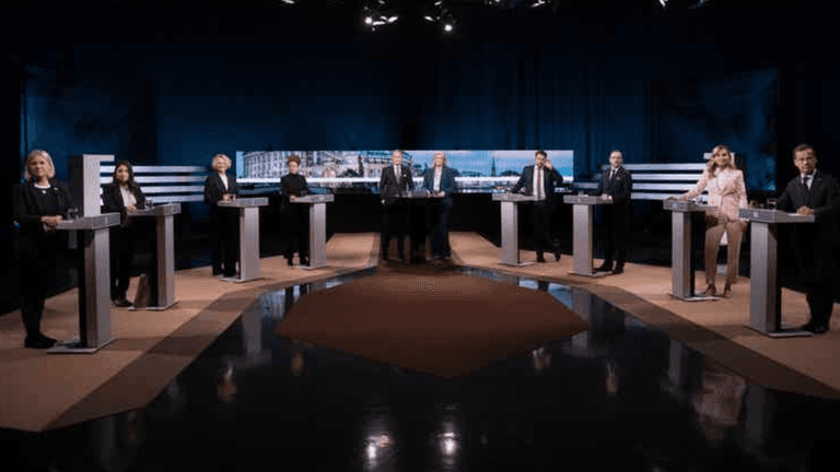 Eight individuals standing at podiums in a debate setting with a dark background